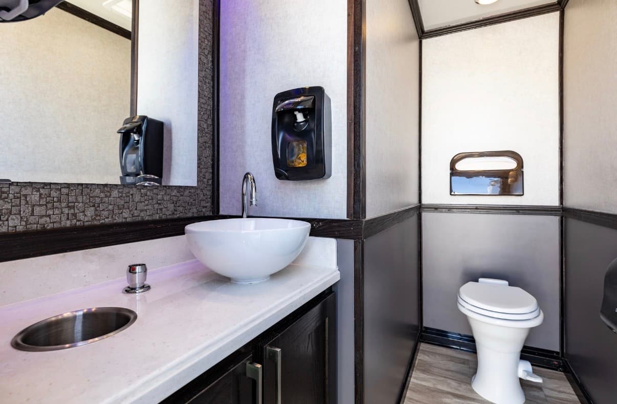 Modern portable restroom interior with white vessel sink, black soap dispenser, and white toilet.